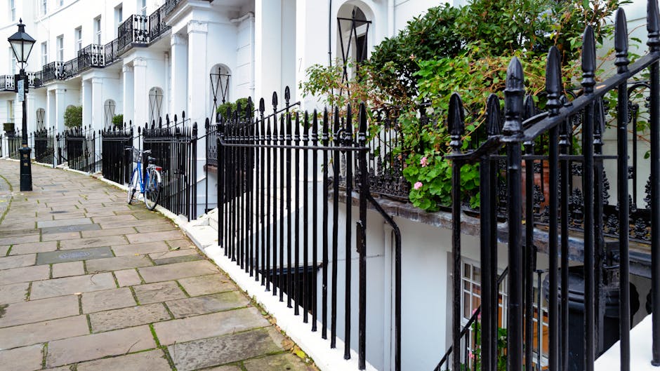 A row of white terraced houses along Brondesbury Road features a paved sidewalk with rectangular stone slabs, some showing weathered textures, leading past black wrought iron railings that enclose sma